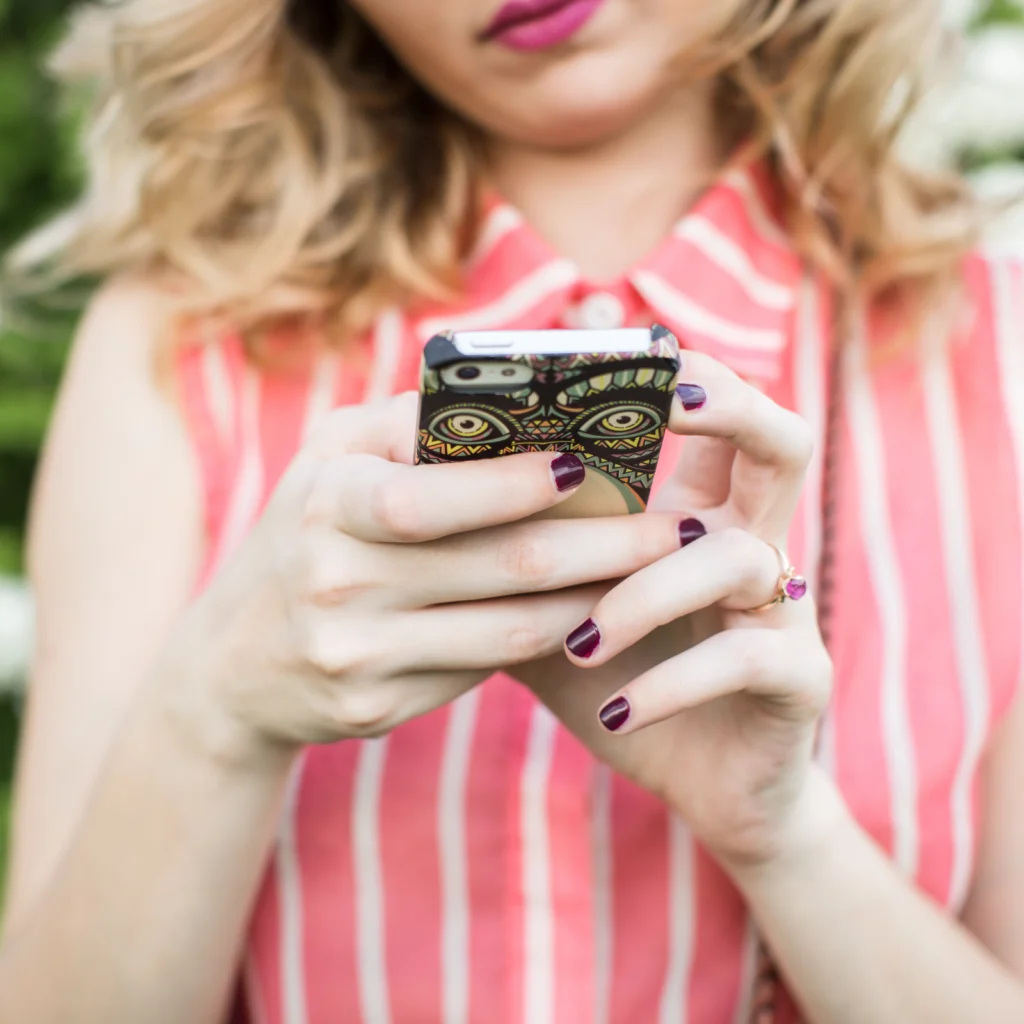 A woman in a pink striped dress uses a phone with an eye-patterned case. She has wavy blonde hair and dark purple nail polish.