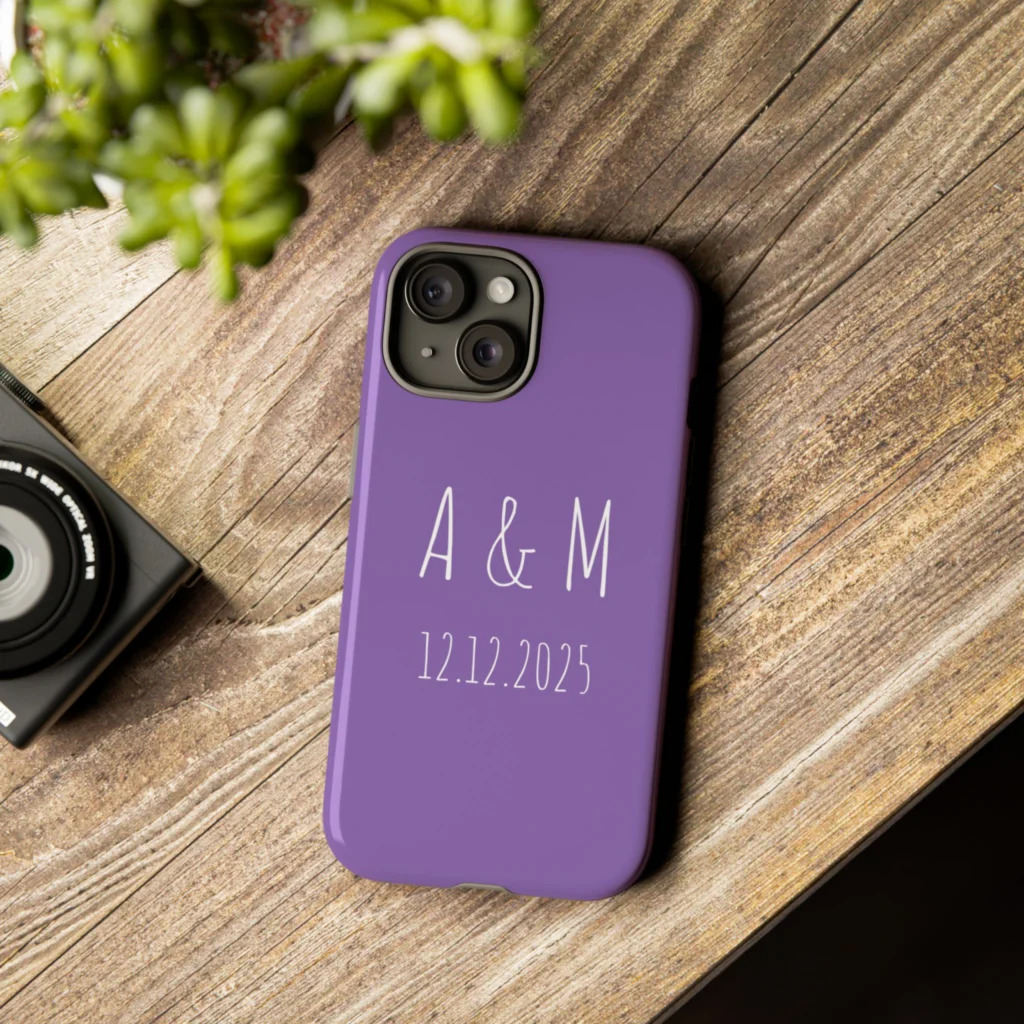 Purple phone case with "A & M 12.12.2025" text, sits on a wooden table beside a camera and potted plant.