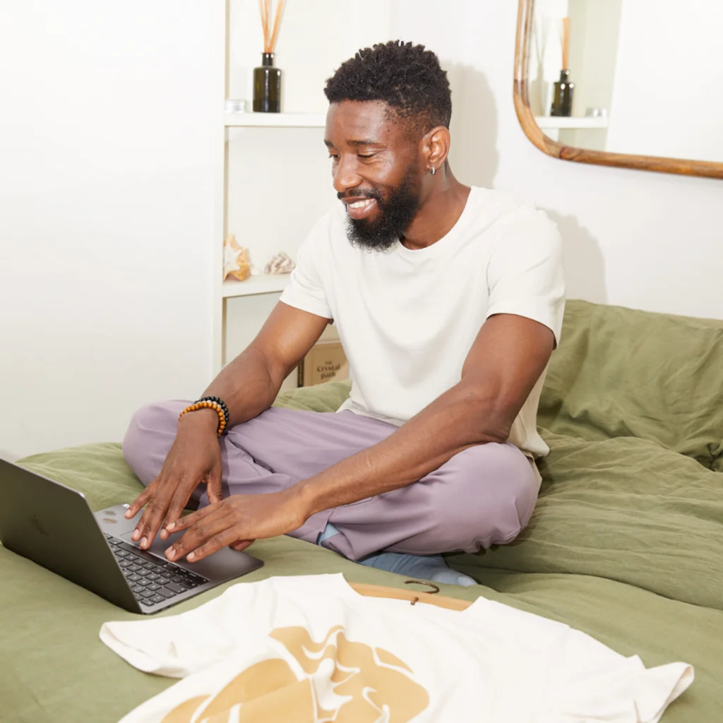A smiling man is sitting on top of a bed with a laptop in front of him. A white t-shirt with a golden print is nearby.