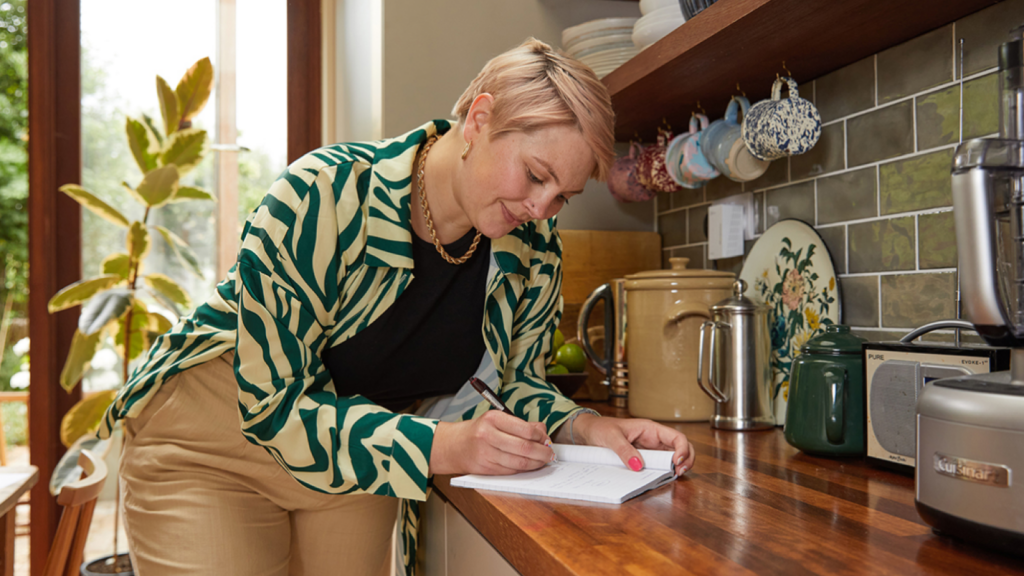 A young woman with short hair leans on a kitchen counter, writing in a notepad. They wear a green and beige striped top.