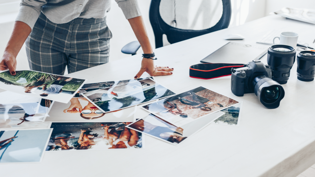 A person in plaid pants arranges printed photos on a white desk with a camera, lenses, and a laptop nearby.