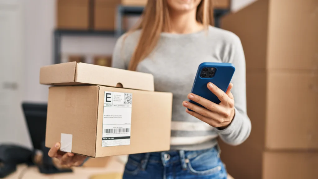A woman holding a cell phone in one hand and a package in the other, smiling while standing indoors.