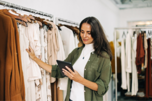 A woman stands in front of clothes, intently looking at a tablet in her hands.