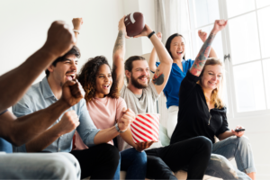 A diverse group of people excitedly watches a sports game indoors. They cheer with arms raised, holding a football and popcorn.