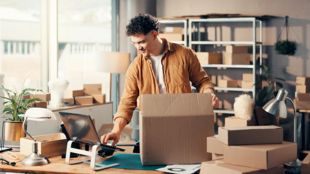 01 How to validate your Etsy business ideas before launching A man in a brown shirt happily packs a large cardboard box in a modern office filled with stacked boxes, a computer, and plants.