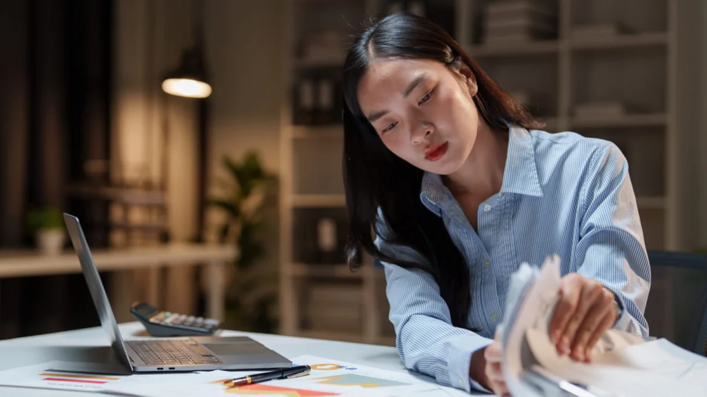 Young woman in a blue shirt working intently at a desk with a laptop. She reviews documents with focus.