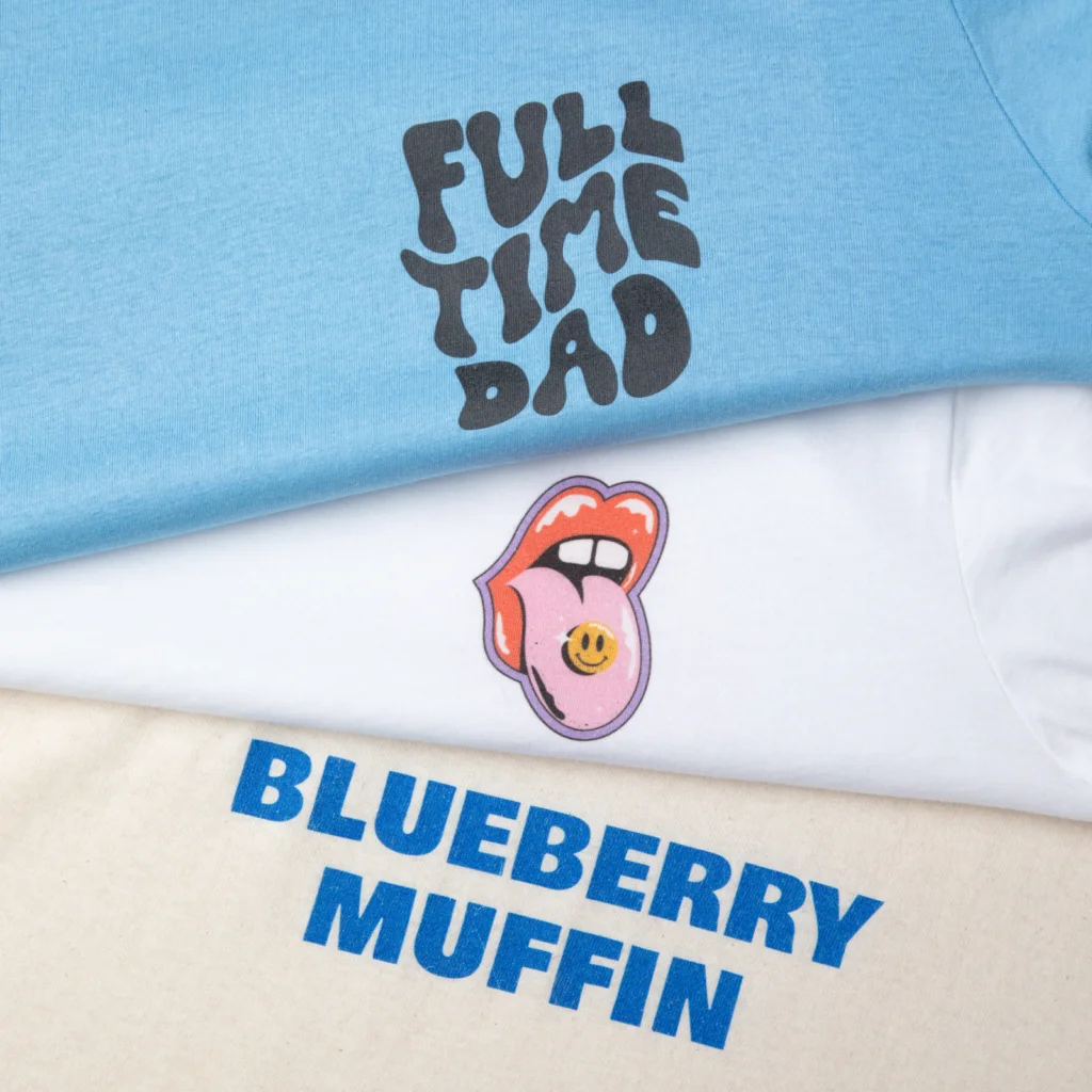 Three t-shirts stacked: the top blue with "Full Time Dad," middle white with a graphic of lips and tongue, bottom beige with "Blueberry Muffin".