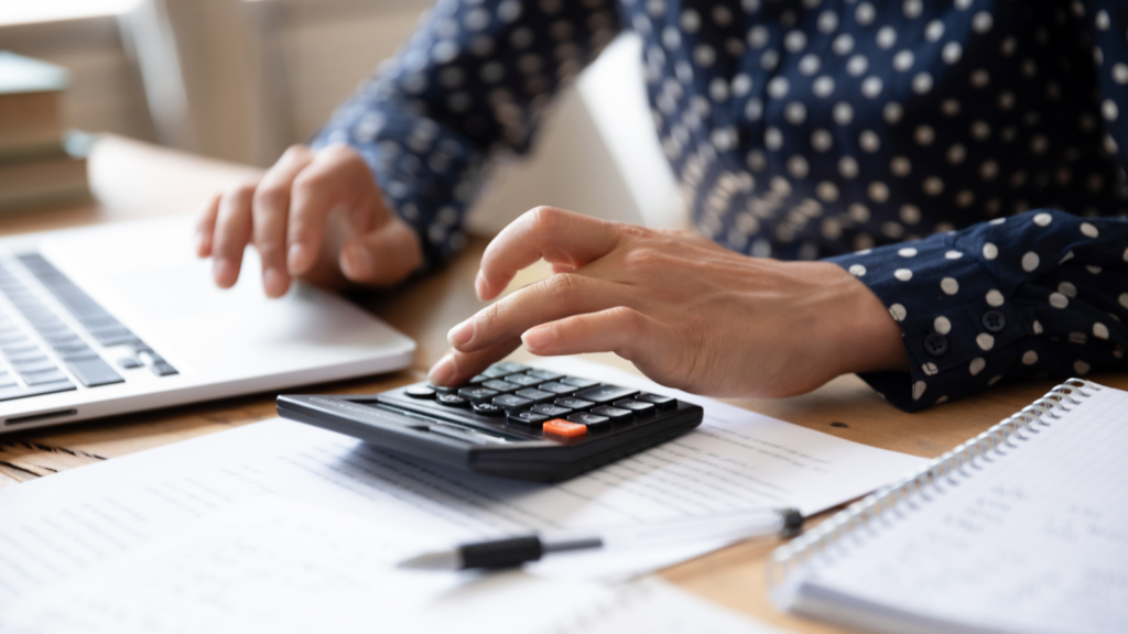 A person in a polka dot shirt uses a calculator and a laptop at a desk. Papers and a notebook with a pen are nearby.