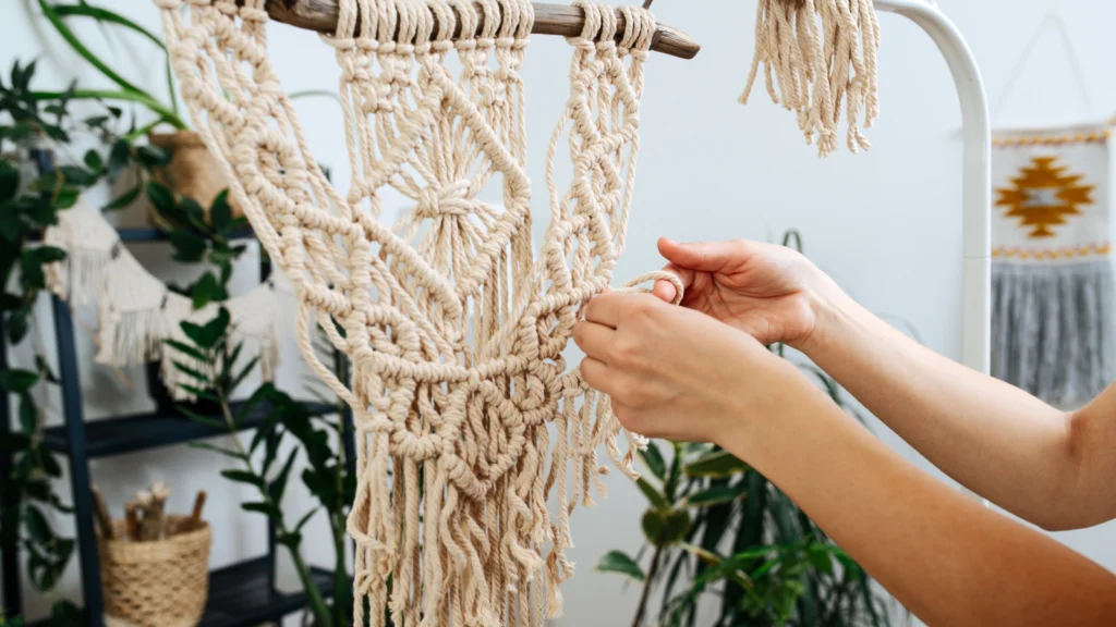 03 Macrame wall art Close-up of hands crafting a macramé wall hanging with intricate patterns. The setting is a bright room with plants.