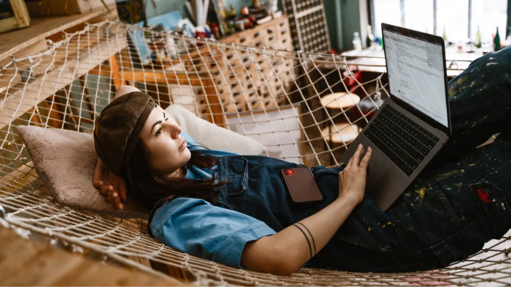 A woman wearing a hat and overalls relaxes on a net hammock indoors, working on a laptop to learn how to make money on Etsy.