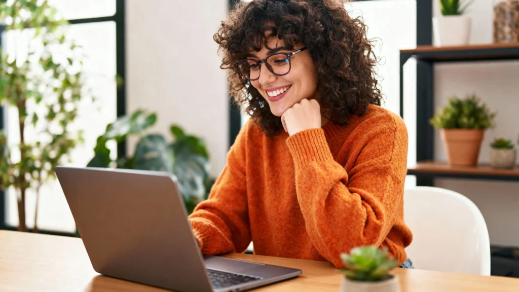 A woman with curly hair and glasses, in an orange sweater, smiles as she uses a laptop at a wooden table. Plant and shelves in the background.