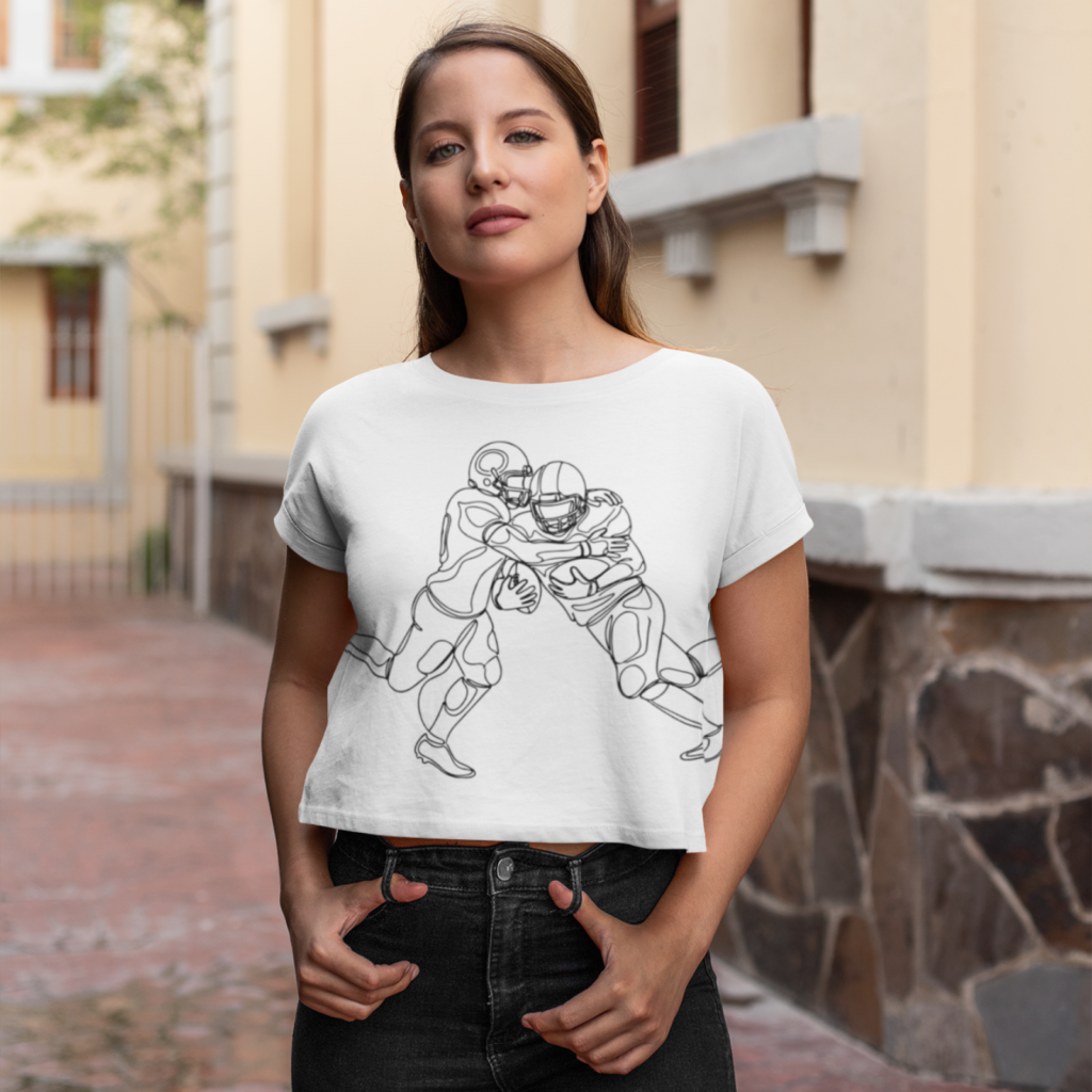 05 Football line art Super Bowl tee A woman wearing a white crop top with a sketch design of two American football players. She stands confidently in an urban setting.