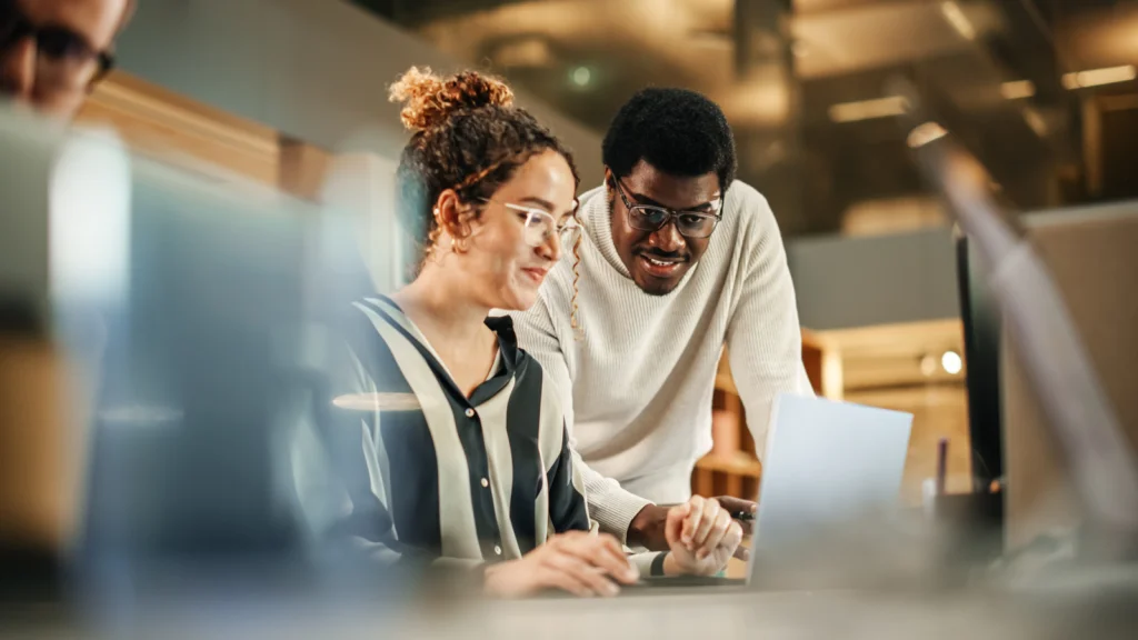 Two people in an office setting, one sitting and the other standing, collaborate over a laptop.