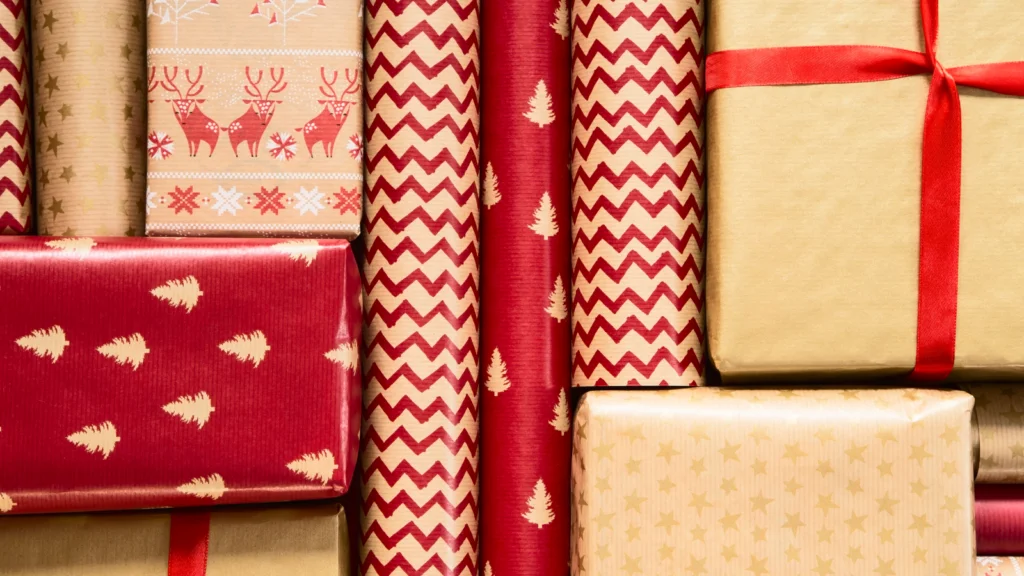 A variety of gift boxes wrapped in red and gold paper, and wrapping paper rolls, featuring festive patterns like reindeer, trees, and zigzags.