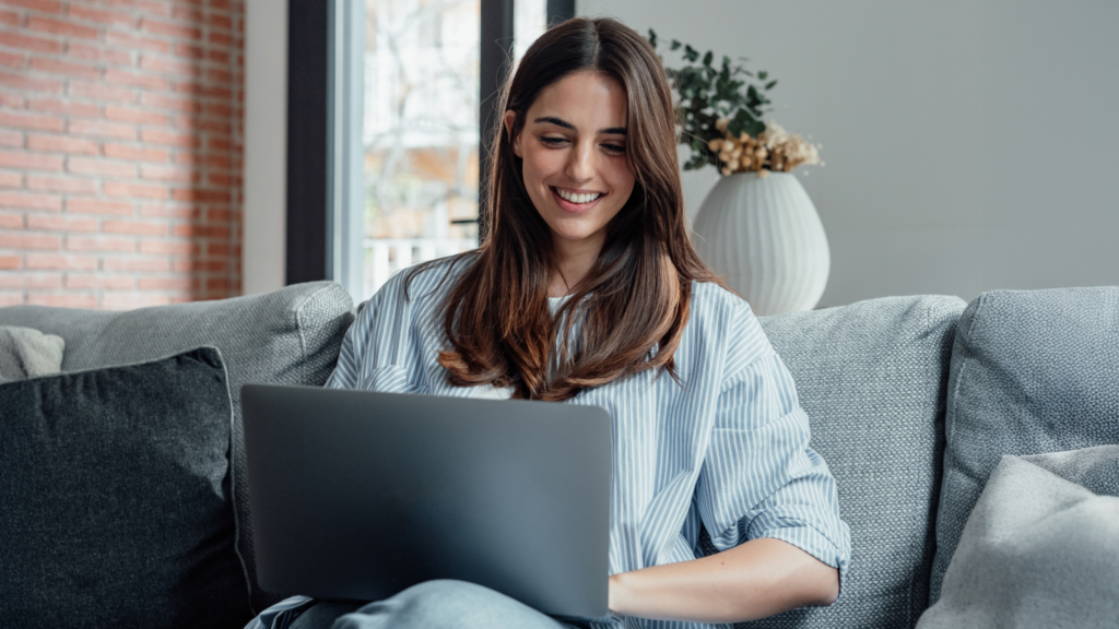 07 How to choose the best idea for an Etsy shop A woman with long brown hair sits on a gray couch, smiling as she searches for Etsy shop ideas on a laptop. A vase with flowers is in the background.