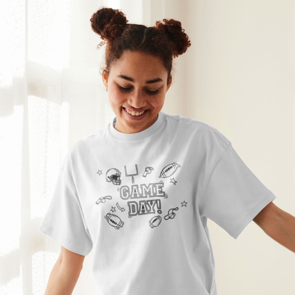 10 Game Day doodle shirt designs A smiling woman wearing a white "Game Day" t-shirt with football elements drawn as doodles, standing by a sunlit window.