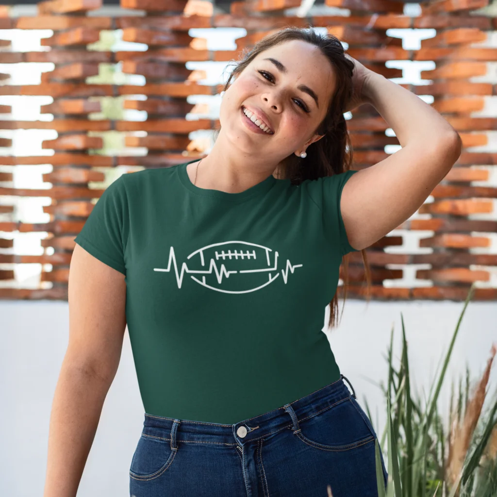 12 Minimal football heartbeat design A smiling woman in a green shirt with a white heartbeat and football design, standing outdoors with a brick wall background.