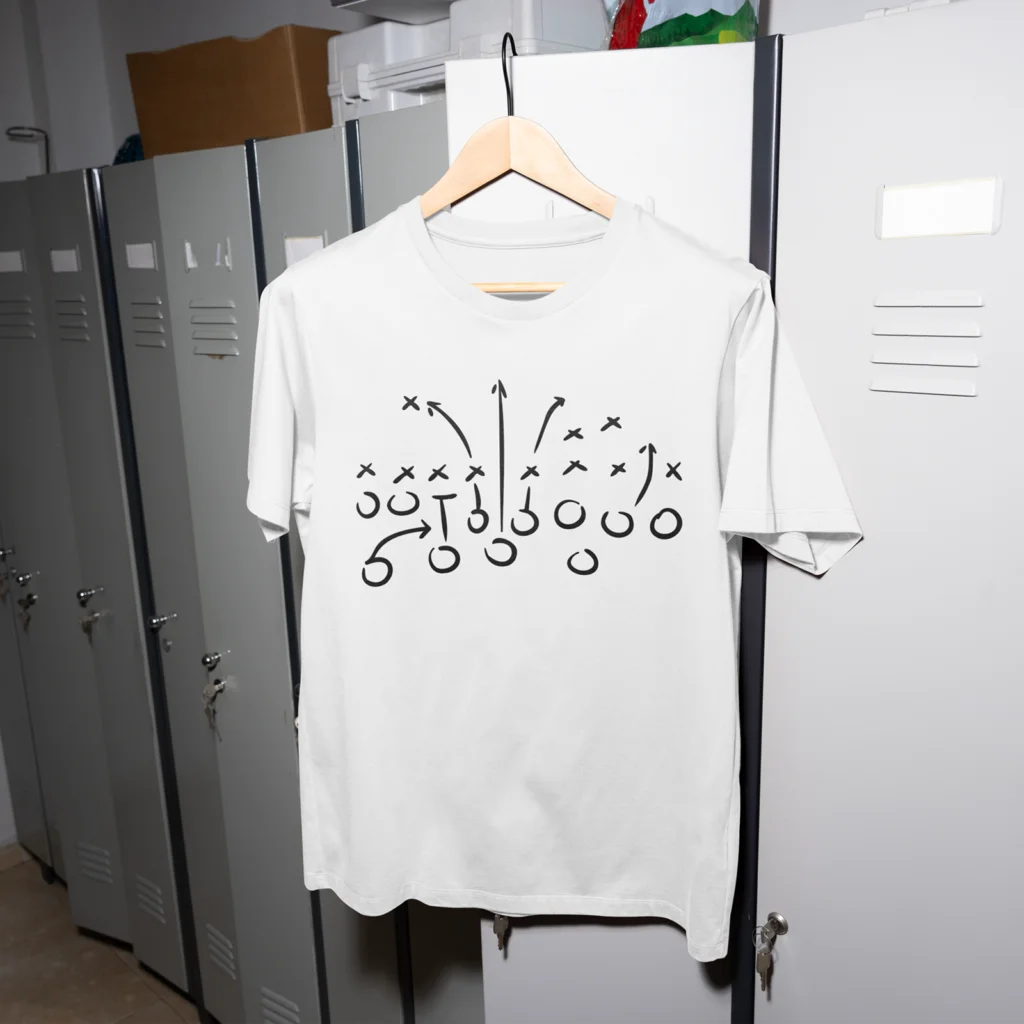 13 Football playbook t shirt A white t-shirt with a black football play diagram, featuring X’s and O’s with arrows, hangs on a wooden hanger in front of gray lockers.