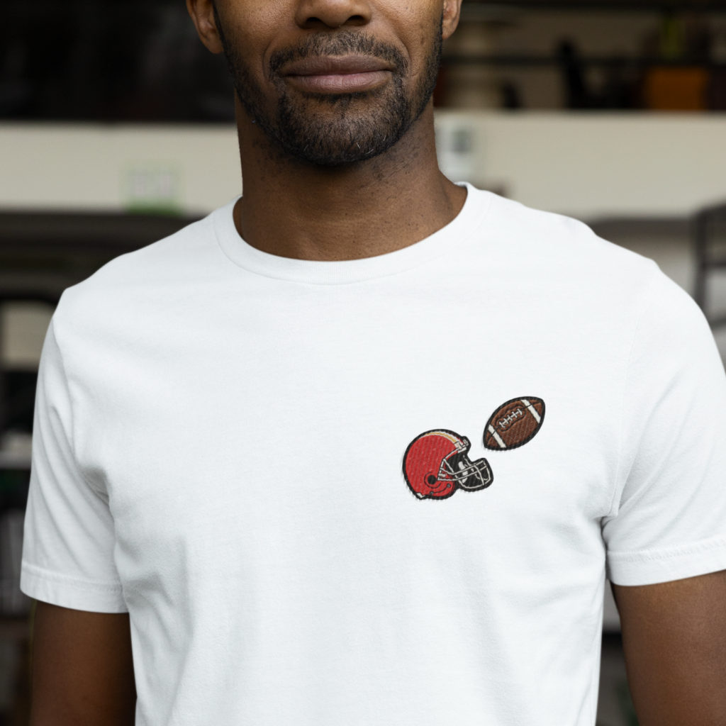 14 Embroidered football themed graphics A man wearing a white t-shirt with a small embroidered red football helmet and a brown football.
