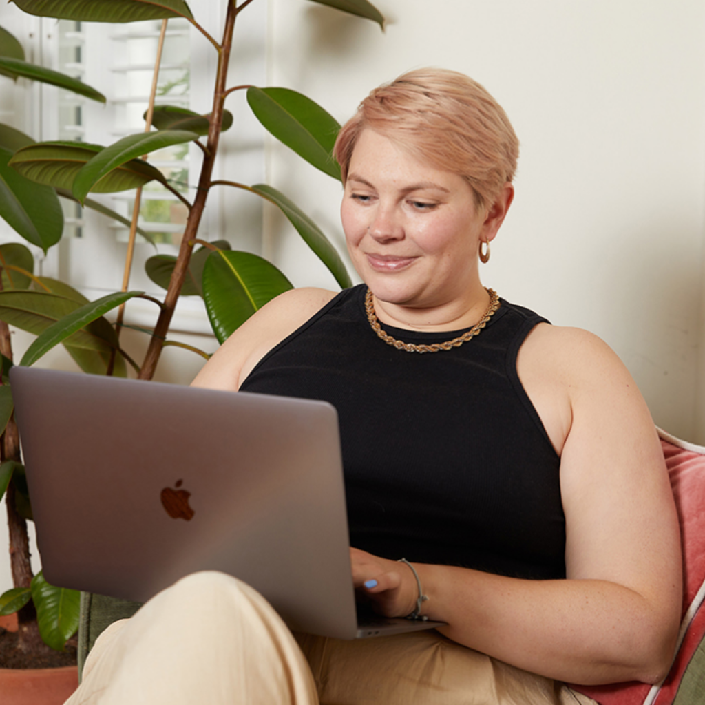 16 How to design Super Bowl shirts using Printifys Product Creator A woman with short hair sits on a sofa, smiling at a laptop in a cozy setting. A large green plant is nearby.