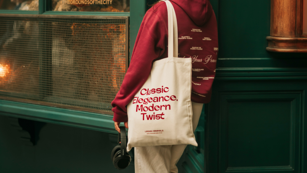 A woman in a red sweatshirt carries a tote bag, standing confidently with a casual demeanor.
