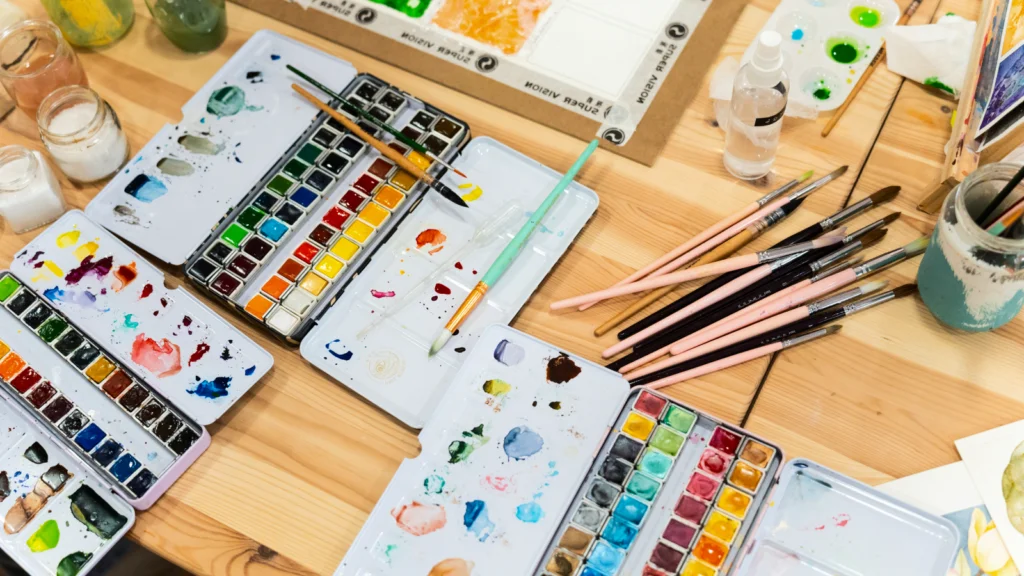 Watercolor painting supplies, including brushes, paints, and paper, arranged neatly on a wooden table.
