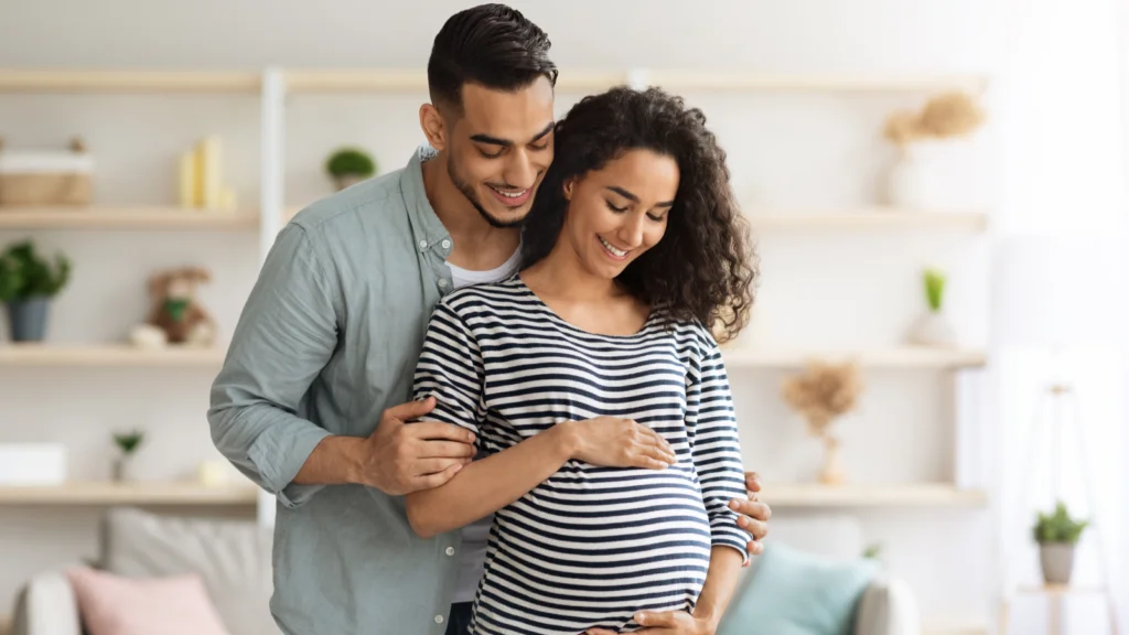 A pregnant woman and her husband stand together in a cozy living room, smiling and embracing the moment.
