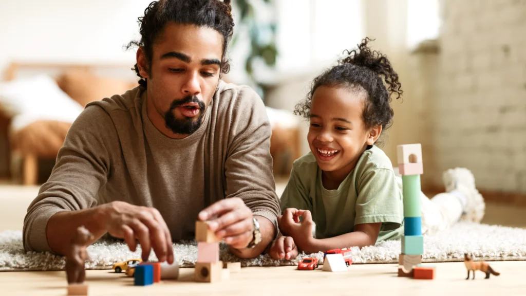 A man and a child joyfully building a structure with colorful toy blocks on a play mat.