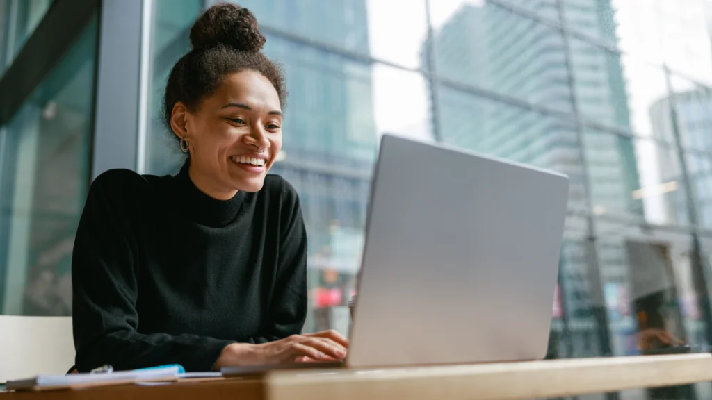 A smiling woman in a black sweater works on a laptop in a modern glass office. Cityscape visible through large windows.