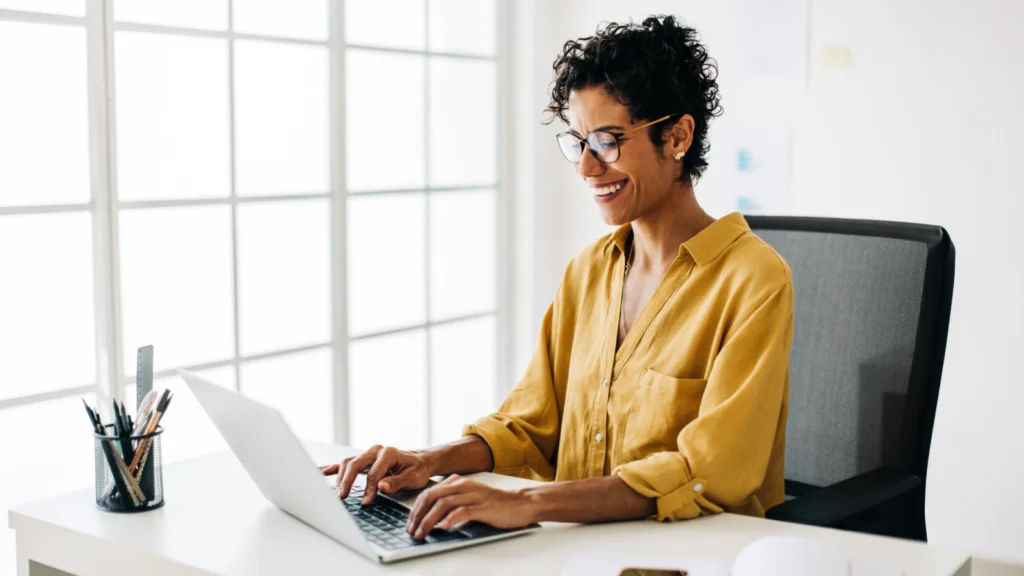 A smiling woman with curly hair and glasses, wearing a yellow shirt, types on a laptop in a bright office.