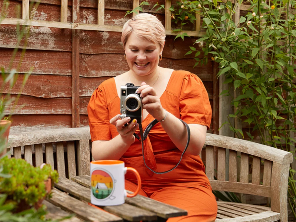 A smiling woman in an orange dress sits on a wooden bench in a garden, taking a photo of a colorful mug using a digital camera.