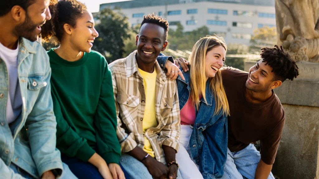 A group of five young adults sits together, laughing and smiling outdoors on a sunny day.