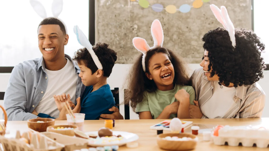 A joyful family of four, wearing bunny ears, sits around a table decorated with Easter crafts and colorful eggs.