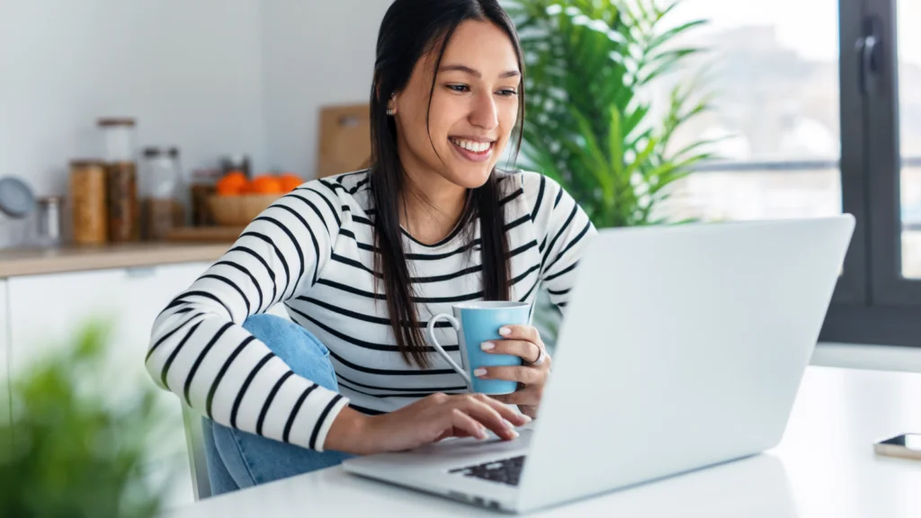 A smiling woman in a striped shirt uses a laptop to learn how to design custom jewelry at a bright kitchen table, holding a blue mug.