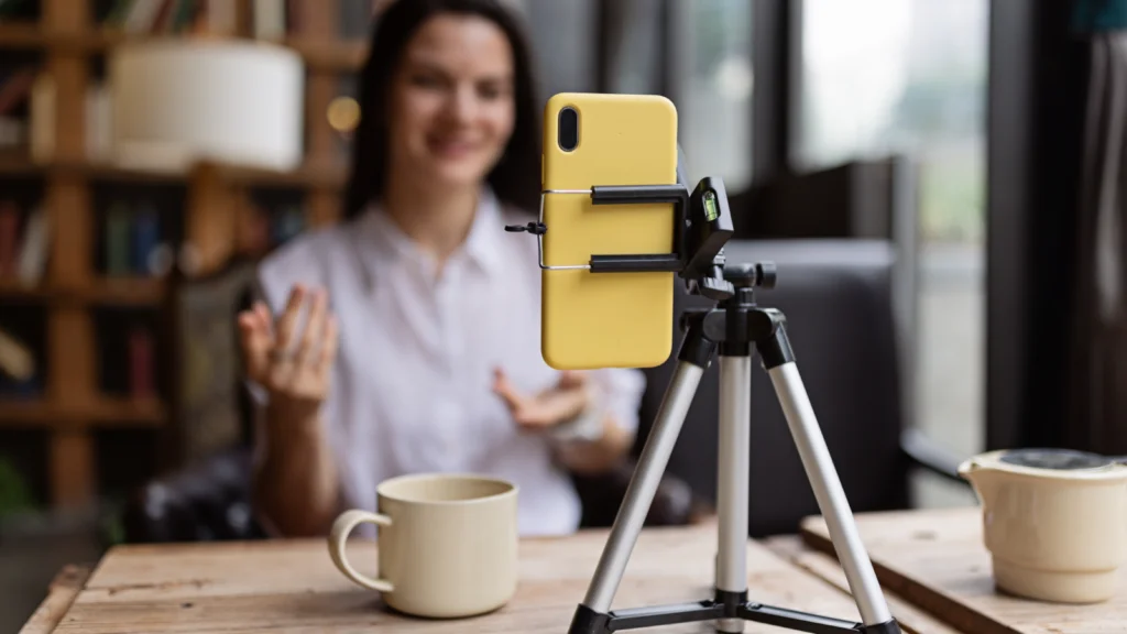 A smiling woman in a cozy room is recording herself with a smartphone on a tripod, set on a wooden table with a coffee cup nearby.