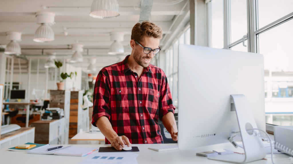 A man in a checkered shirt is focused on working at his computer, surrounded by a tidy workspace.