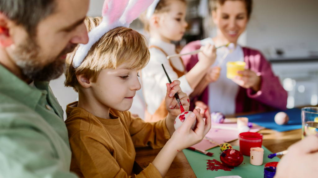 A young boy with bunny ears paints an Easter egg, surrounded by family.