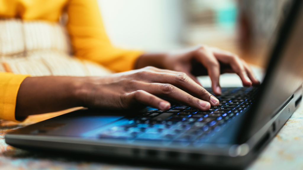 Hands typing on a laptop keyboard, with a person wearing a yellow top in the background.