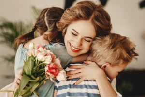 A woman embraces two children, surrounded by colorful flowers, conveying warmth and affection in a joyful moment.