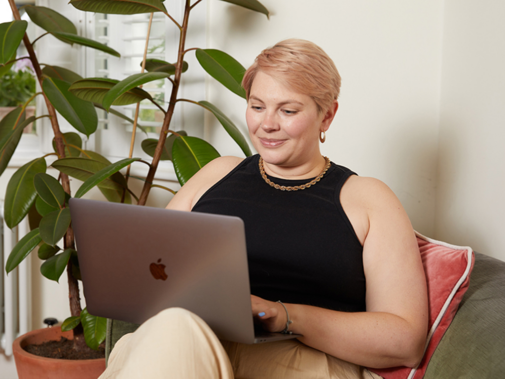 A woman with short blond hair sits on a couch using a laptop, searching for side hustles for women. A large green plant is in the background.