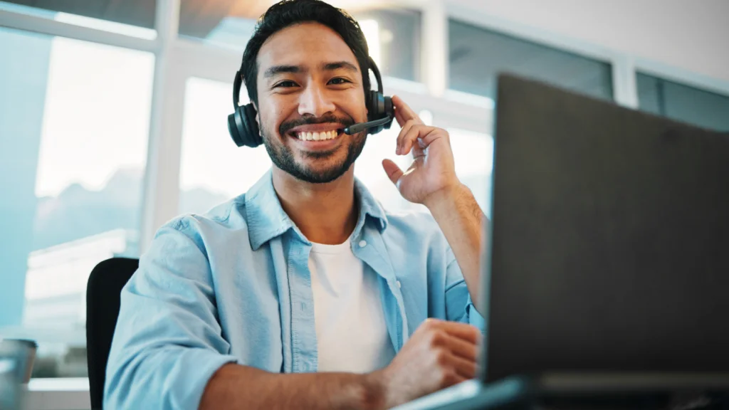 A smiling man wearing a headset sits at a desk with a laptop, appearing engaged. Bright office space with large windows.