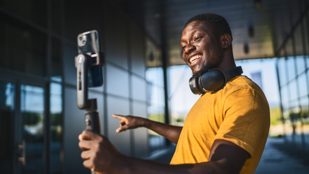 A man in a yellow shirt smiles and points at a smartphone on a tripod, filming himself, wearing headphones around his neck.