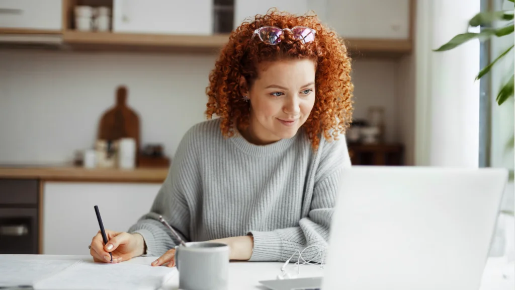 A woman with curly red hair is smiling at a laptop while taking notes in a cozy kitchen.