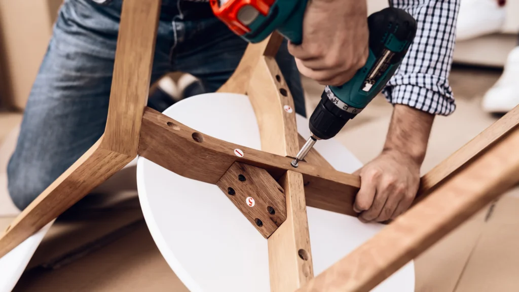 A man uses a power drill to assemble a wooden table with a white surface. The focus is on his hands and the tool.