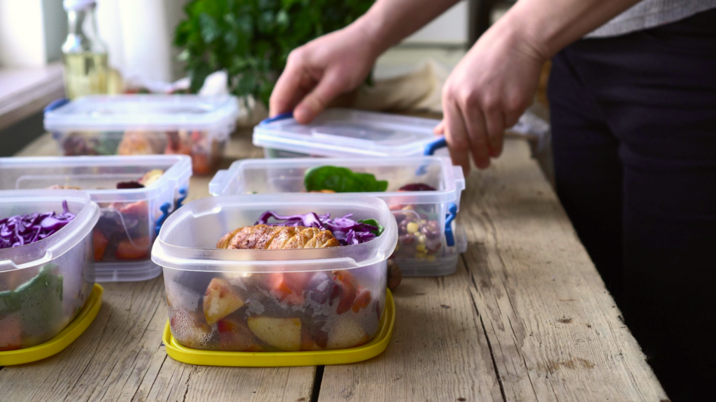 Hands sealing food containers with colorful salads and roasted vegetables on a wooden table.