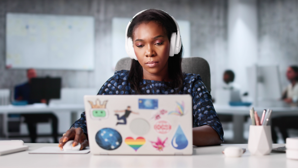 A focused woman wearing headphones works on a sticker-covered laptop in a modern, open-plan office.