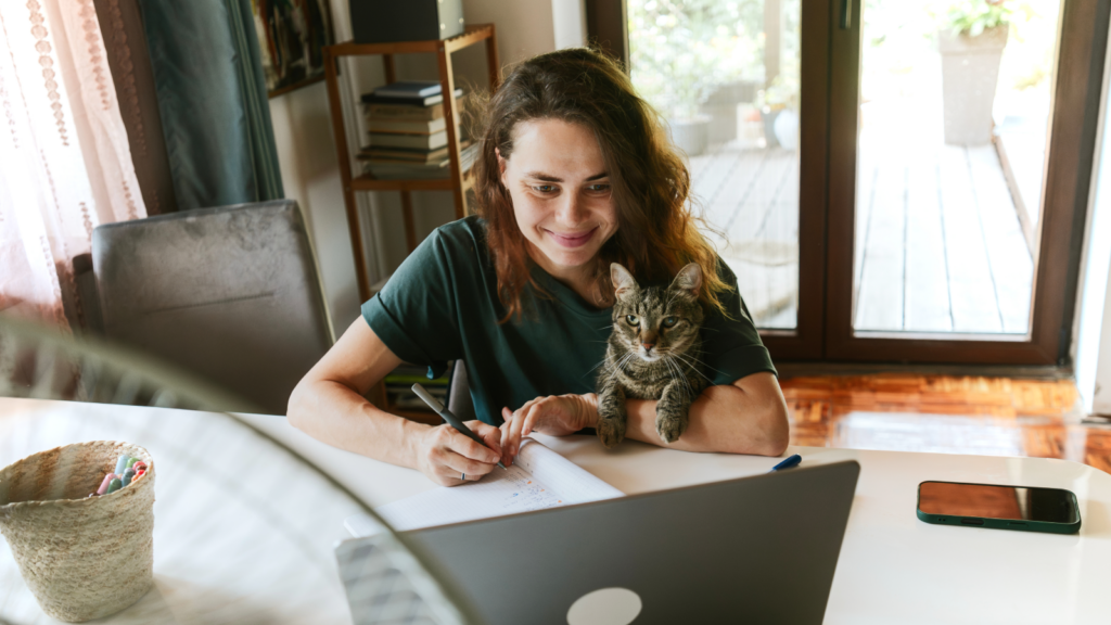 A woman smiling while working on a laptop at home, with a cat by her side, a phone, and a basket on the table.