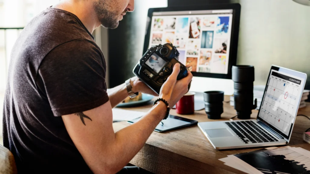 A man at a desk holds a camera, reviewing photos. An open laptop and monitor display images.