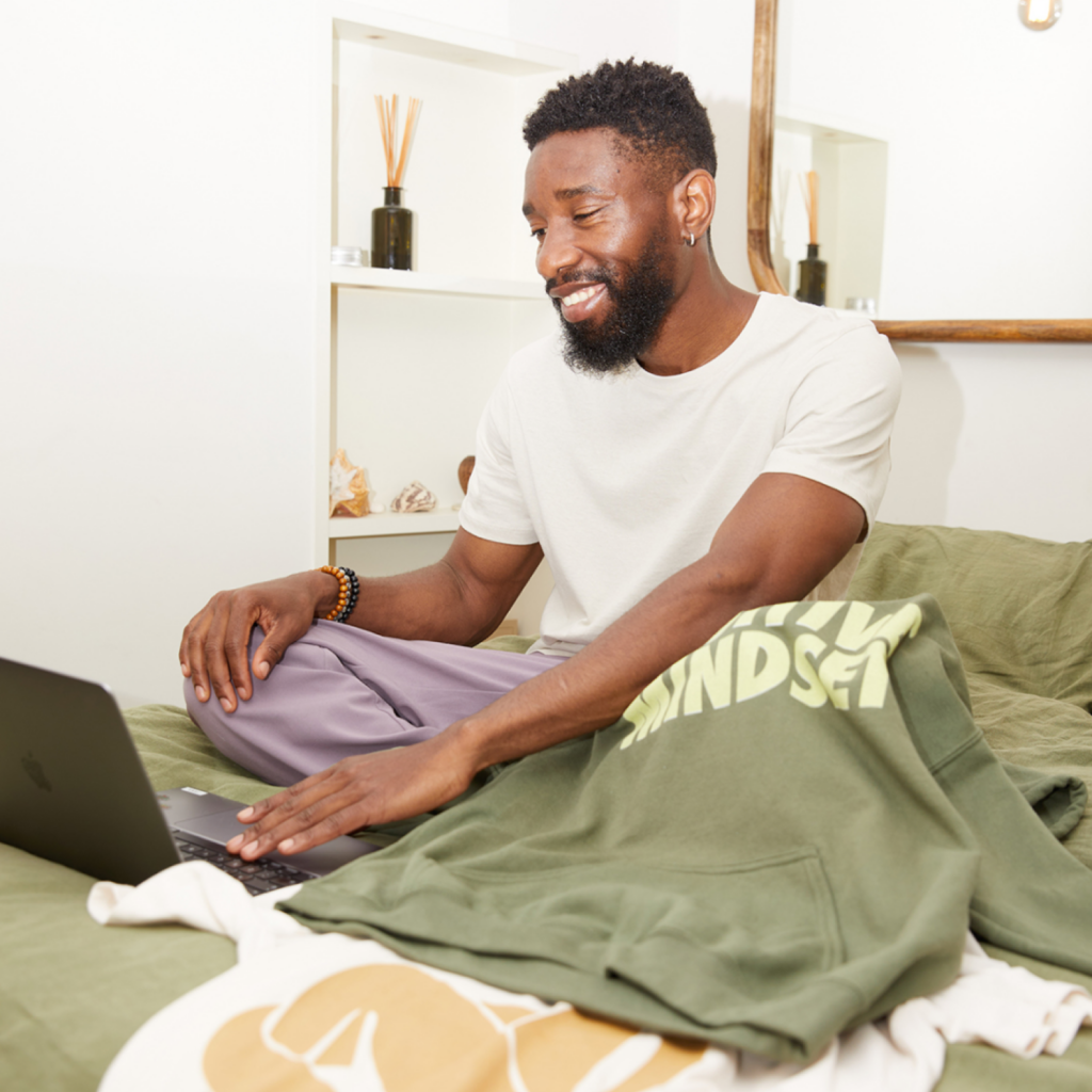 A man smiles while using a laptop, seated on a bed, with a green hoodie with a text print.