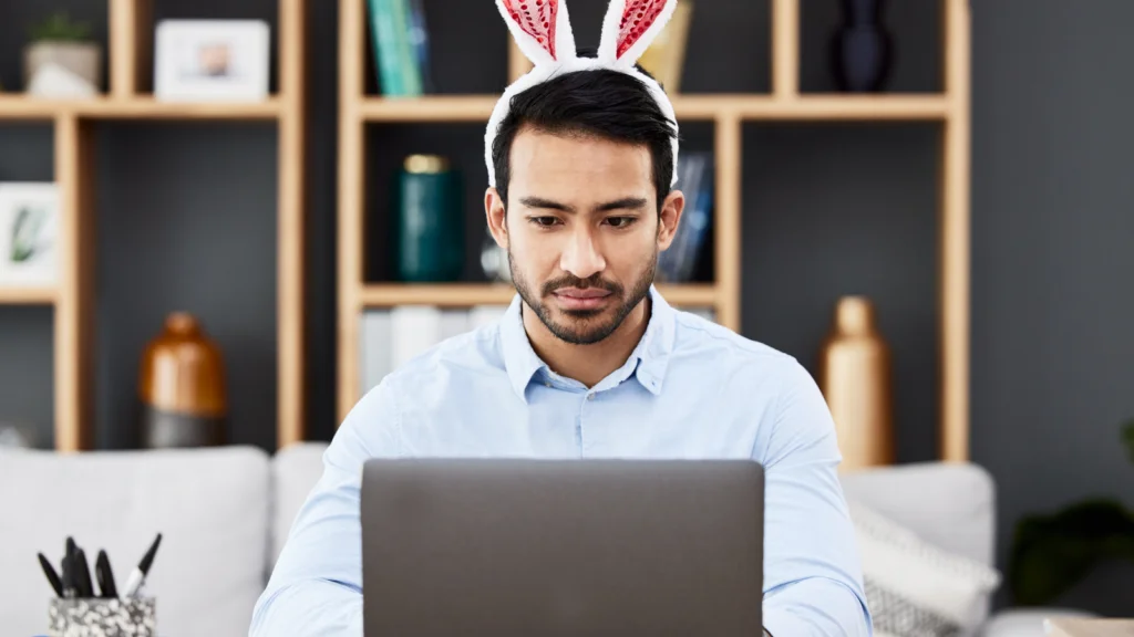 A man wearing bunny ears focuses intently on a laptop, seated in a modern living room with shelves and decor.